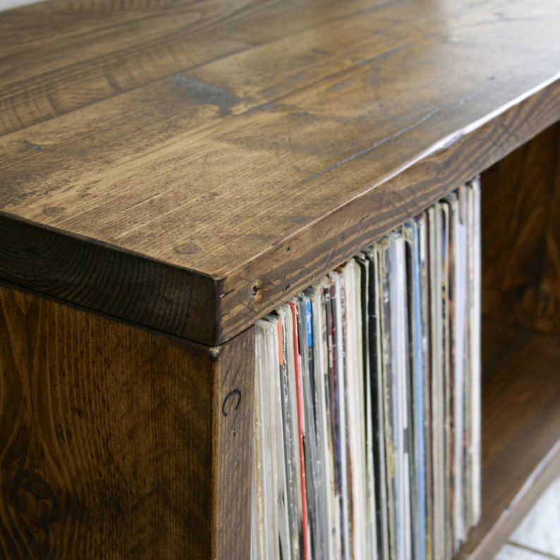 May include: A dark brown wooden record player stand with a shelf for storing vinyl records. The stand has a natural wood finish and is made of solid wood.
