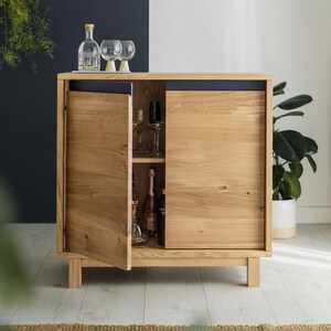May include: A light oak cabinet with two doors, one ajar, displaying glassware and bottles. The cabinet rests on short legs. Two wine glasses and a bottle are on top. The background is a mix of dark blue and white.
