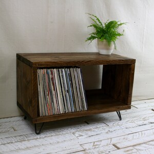 May include: A rustic wooden record player stand with a shelf for storing vinyl records. The stand has a dark brown stain and is supported by hairpin legs. A potted plant sits on top of the stand.