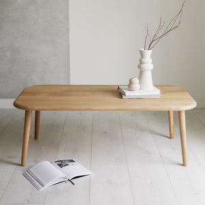 May include: A light wood coffee table with rounded edges and four legs. The table is decorated with a white vase with a branch of twigs, a white ceramic ornament, and a stack of books.