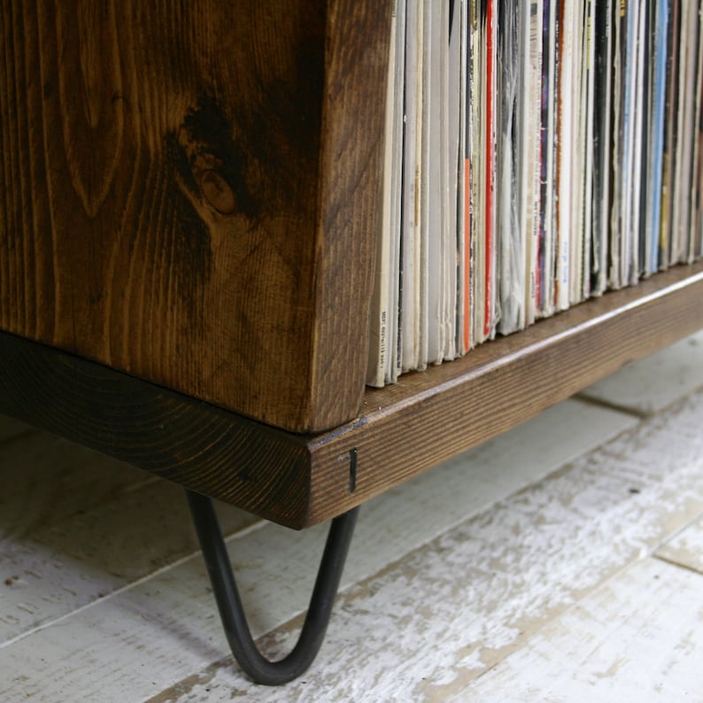 May include: A dark brown wooden record player stand with black hairpin legs. The stand is filled with vinyl records.