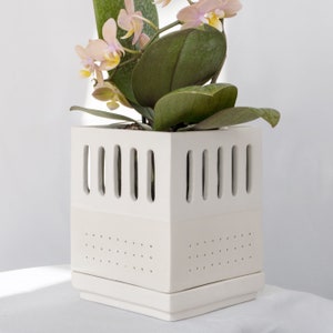 Può includere: Un vaso di ceramica bianco con forma quadrata e design geometrico. Il vaso ha una serie di piccoli fori sul fondo e aperture più grandi sui lati. Una pianta di orchidea rosa sta crescendo nel vaso.