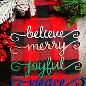 May include: Four wooden Christmas signs with a red and black buffalo plaid background. The signs read "believe", "merry", "joyful", and "peace".