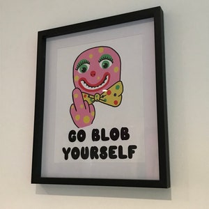 May include: A framed print featuring a pink cartoon character giving the middle finger. The character has a wide smile, green eyes, and a yellow bow tie with polka dots. The text below the character reads "Go Blob Yourself."
