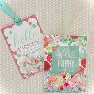 May include: Two rectangular gift tags with shaker elements. The left tag is pink with the text "hello THERE I'VE MISSED YOU FRIEND" and a light blue ribbon. The right tag features a floral pattern, the text "HAPPY today", and a sheer ribbon.