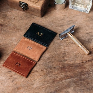 May include: Three leather card holders with initials 'AZ', 'FP', and 'RS' embossed in gold on a wooden surface with a shaving brush, a safety razor, and a small wooden box.