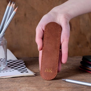May include: A brown leather pencil case with the initials "AK" embossed in gold. The case is held in a hand.