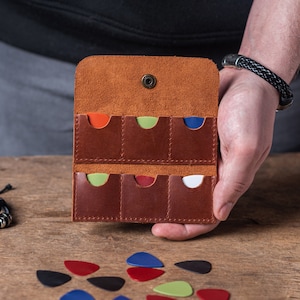 Puede incluir: Un porta-púas de cuero marrón con seis ranuras para púas. El porta-púas está abierto y muestra una variedad de púas de colores en el interior. El porta-púas está siendo sostenido en una mano.