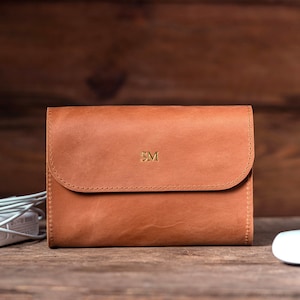 May include: A brown leather laptop case with a flap closure and gold initials "SM". The case is on a wooden surface with a white mouse and a white charging cable.