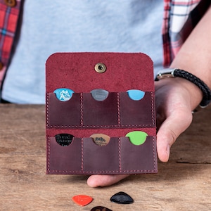 May include: A burgundy leather guitar pick holder with six slots for storing picks. The holder is open and shows a variety of picks in different colors and designs.