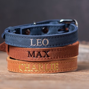 May include: Three leather dog collars stacked on top of each other. The top collar is blue with the name "LEO" embossed in silver. The middle collar is brown with the name "MAX" embossed in black. The bottom collar is brown with the name "CHARLIE" embossed in gold.