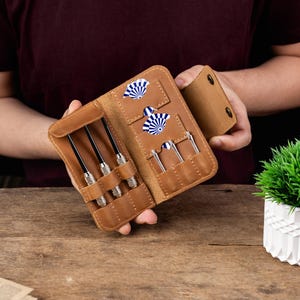 May include: A tan leather dart wallet featuring dedicated slots for three dart barrels and two sets of blue and white patterned dart flights. The folding case includes a secure snap closure and internal pockets to organise dart components.