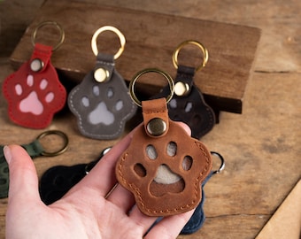 May include: Leather paw print keychains in various colors, including brown, red, gray, black, and green. Each keychain features a gold-colored ring and a metal rivet. The keychains are designed with a paw print cutout.