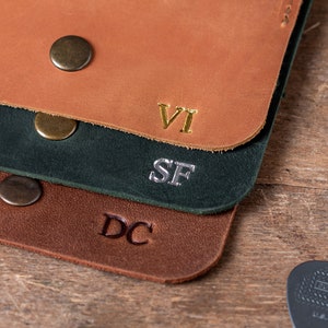 May include: Three leather straps with different coloured stitching and metal rivets. The straps are stacked on top of each other. The top strap is brown with gold lettering that says "VI". The middle strap is green with silver lettering that says "SF". The bottom strap is brown with brown lettering that says "DC".