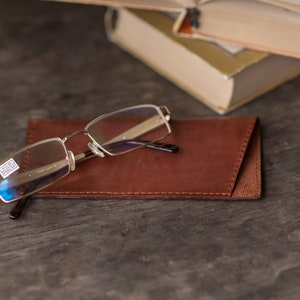 May include: A pair of silver-rimmed glasses with clear lenses resting on a brown leather glasses case. The glasses case has a stitched edge.