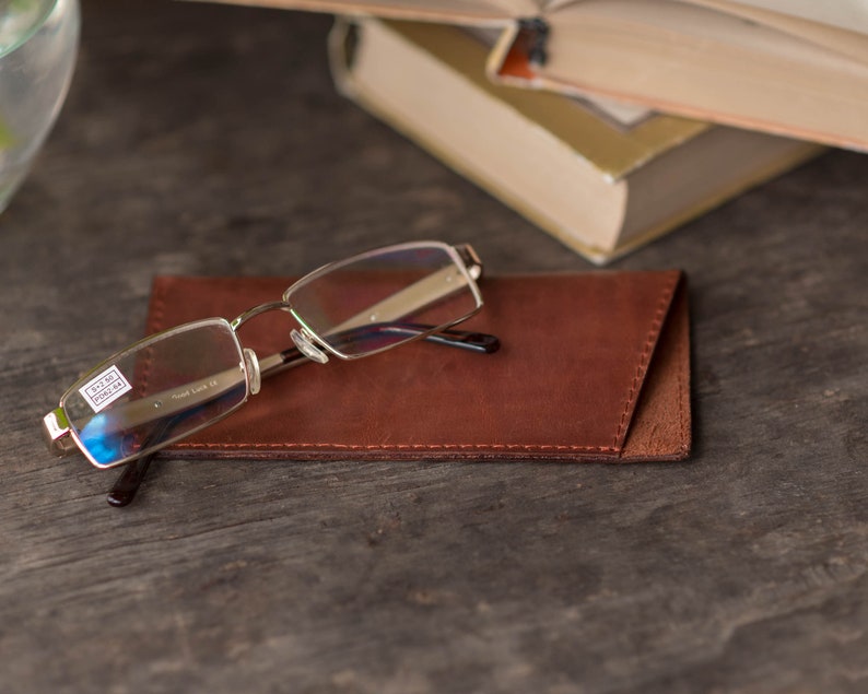 May include: A pair of silver-rimmed glasses with clear lenses resting on a brown leather glasses case. The glasses case has a stitched edge.