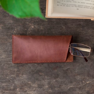 May include: A brown leather glasses case with a flap closure. The case is lying on a wooden surface with a pair of glasses resting on top of it.