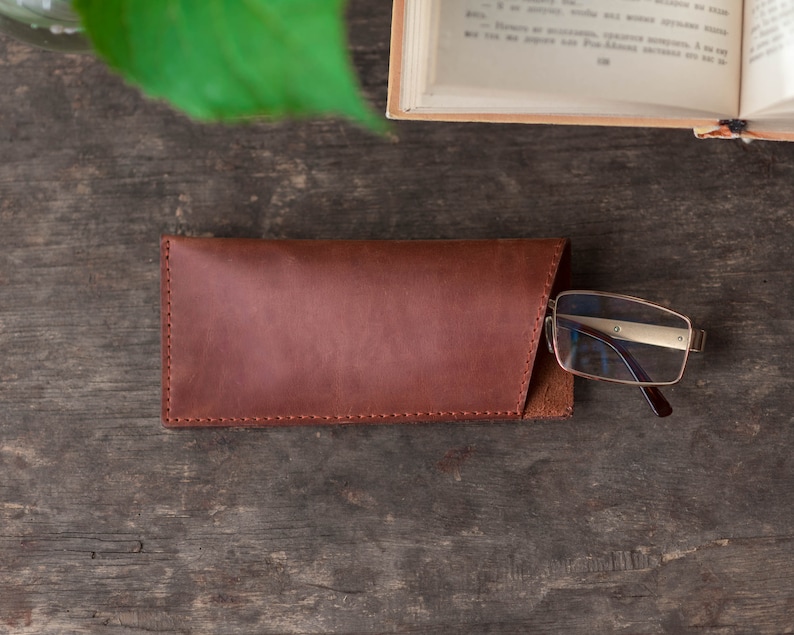 May include: A brown leather glasses case with a flap closure. The case is lying on a wooden surface with a pair of glasses resting on top of it.
