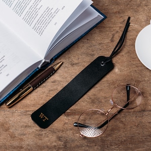 May include: A black leather bookmark with the initials "WT" embossed on it. The bookmark is lying on a wooden surface with a book, a pen, a cup of coffee, and a pair of round glasses.