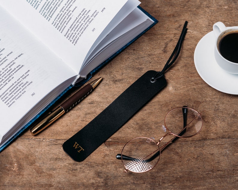 May include: A black leather bookmark with the initials "WT" embossed on it. The bookmark is lying on a wooden surface with a book, a pen, a cup of coffee, and a pair of round glasses.