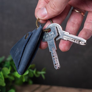 以下が含まれることがあります： 三角形の形状でスナップ留め具が付いた青い革製のキーホルダー。キーホルダーは真鍮製のキーリングにキーセットが取り付けられています。