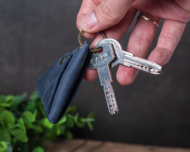 以下が含まれることがあります： 三角形の形状でスナップ留め具が付いた青い革製のキーホルダー。キーホルダーは真鍮製のキーリングにキーセットが取り付けられています。