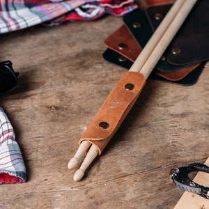 May include: A brown leather drumstick holder with two wooden drumsticks inside. The holder has a brass snap closure and the initials "K.V." are embossed on the front.