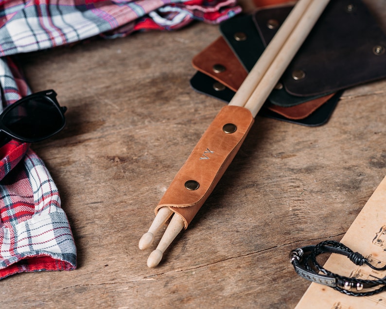 May include: A brown leather drumstick holder with two wooden drumsticks inside. The holder has a brass snap closure and the initials "K.V." are embossed on the front.