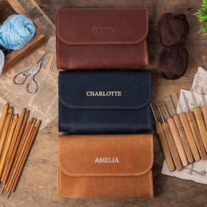 May include: Three leather cases with the names Sophia, Charlotte, and Amelia embossed on them. The cases are closed and are arranged vertically on a wooden surface. A set of wooden crochet hooks is on the right side of the image.