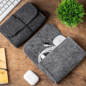 May include: Two gray felt pouches, one open and one closed, with a keyboard and a small green plant in the background. The open pouch contains a white mouse and charging cables. A pair of glasses and a notepad are also visible.