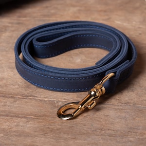 May include: A dark blue leather dog leash with a gold metal clasp. The leash is folded in a circle and the clasp is visible in the foreground.