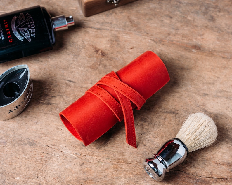 May include: A red leather roll with a white stitching detail, a silver shaving brush with white bristles, and a black bottle with the text "Victory Limited Edition" on the label.