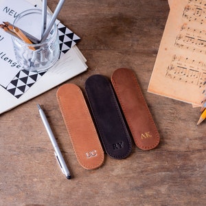 May include: Three brown leather pencil cases with initials "UC", "RY", and "AK" embossed in gold. The cases are on a wooden surface.