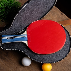 May include: A red ping pong paddle with a blue and white handle rests inside a gray zippered case. A white and a yellow ping pong ball are next to the case on a wooden surface. The paddle has the brand name "DONIC" on the handle.