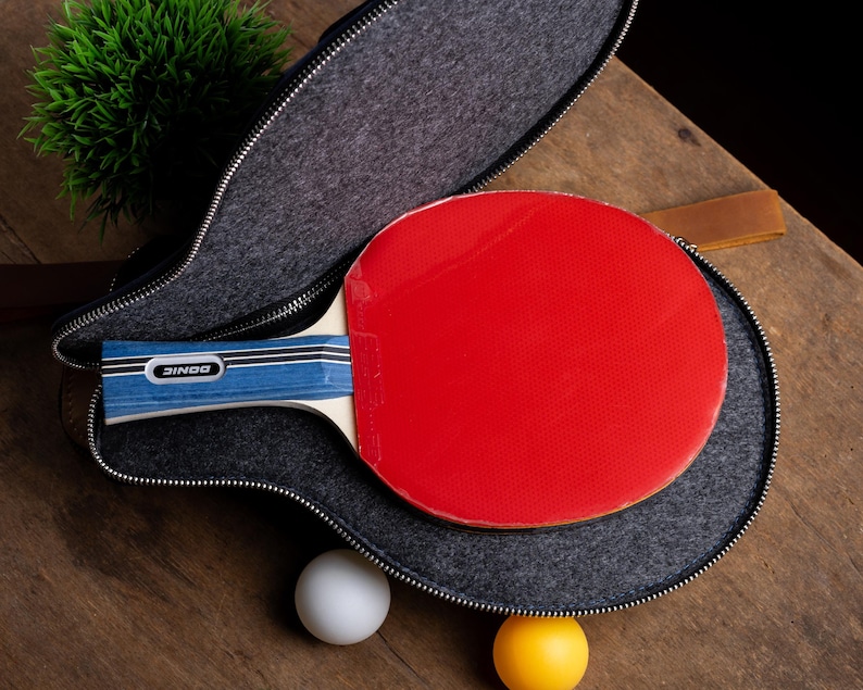 May include: A red ping pong paddle with a blue and white handle rests inside a gray zippered case. A white and a yellow ping pong ball are next to the case on a wooden surface. The paddle has the brand name "DONIC" on the handle.