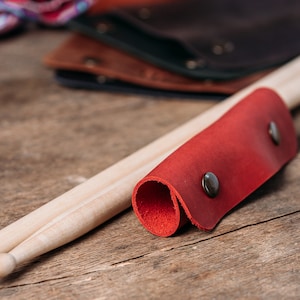 May include: A red leather drumstick holder with two metal poppers. The holder is wrapped around two wooden drumsticks.