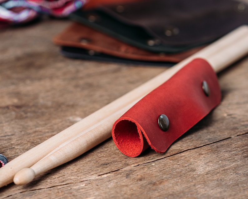 May include: A red leather drumstick holder with two metal poppers. The holder is wrapped around two wooden drumsticks.