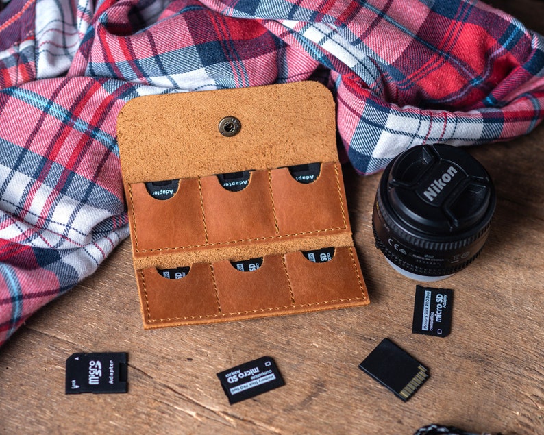 May include: A brown leather case with six slots for memory cards. The case is open and the slots are visible. There is a camera lens and a memory card on the table next to the case.