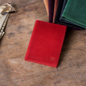 May include: A red leather wallet with the initials "SF" embossed on the front. The wallet is lying on a wooden surface.