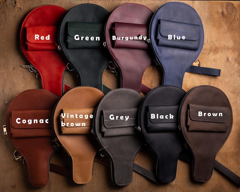 May include: A collection of leather bags in various colors, including red, green, burgundy, blue, cognac, vintage brown, grey, black, and brown. Each bag has a unique shape with a small front pocket and a zipper closure.