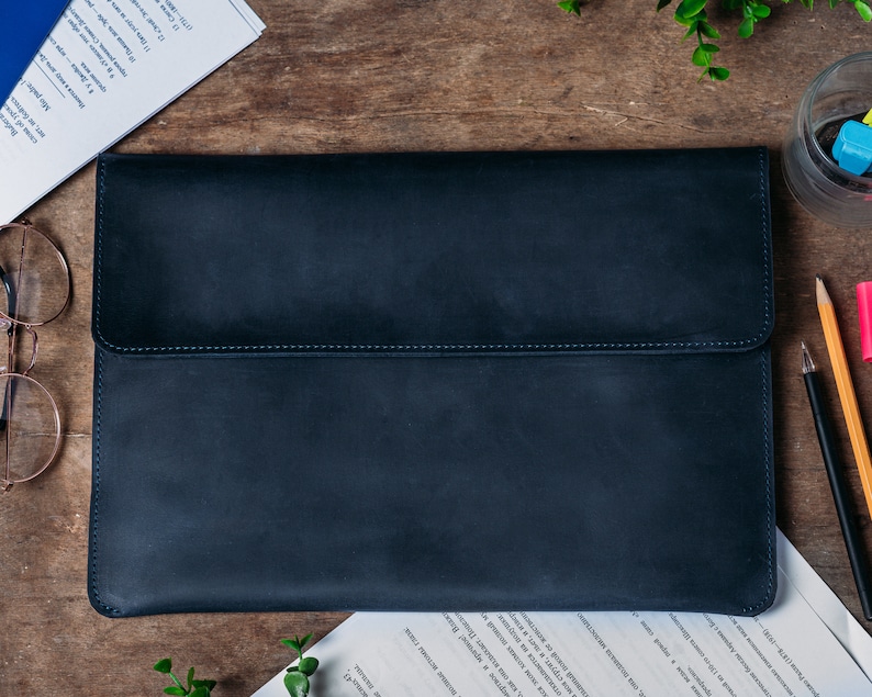 May include: A black leather laptop sleeve with a flap closure. The sleeve is on a wooden surface.
