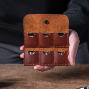 May include: A brown leather case with six slots for memory cards. The case is open and the slots are empty.