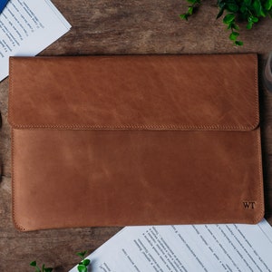 May include: A brown leather laptop sleeve with a flap closure and the initials "WT" embossed on the front.