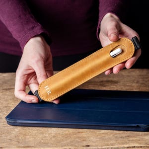 May include: A tan leather stylus holder with the initials "H.R." is held above a dark blue tablet case. The stylus holder has a black strap and a white stylus is visible inside. The background is a wooden surface.