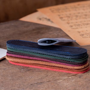 May include: A stack of eight leather pen sleeves in various colours, including blue, green, brown, and red. The sleeves are all closed and stacked on top of each other. A black pen is inside the top sleeve.