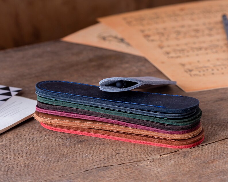May include: A stack of eight leather pen sleeves in various colours, including blue, green, brown, and red. The sleeves are all closed and stacked on top of each other. A black pen is inside the top sleeve.