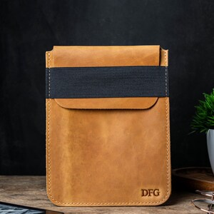 May include: A tan leather tablet sleeve with a black elastic band closure. The sleeve has a flap closure and is embossed with the initials "DFG" in the bottom right corner. The leather has a distressed finish.