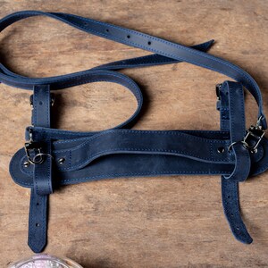 May include: A dark blue leather camera strap with adjustable straps and metal hardware. The strap is designed to be worn over the shoulder.