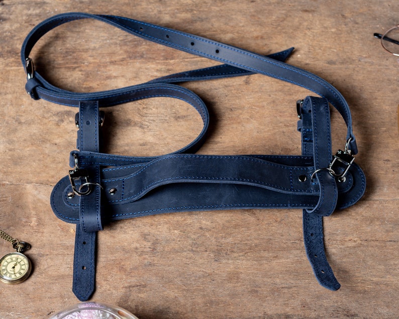 May include: A dark blue leather camera strap with adjustable straps and metal hardware. The strap is designed to be worn over the shoulder.