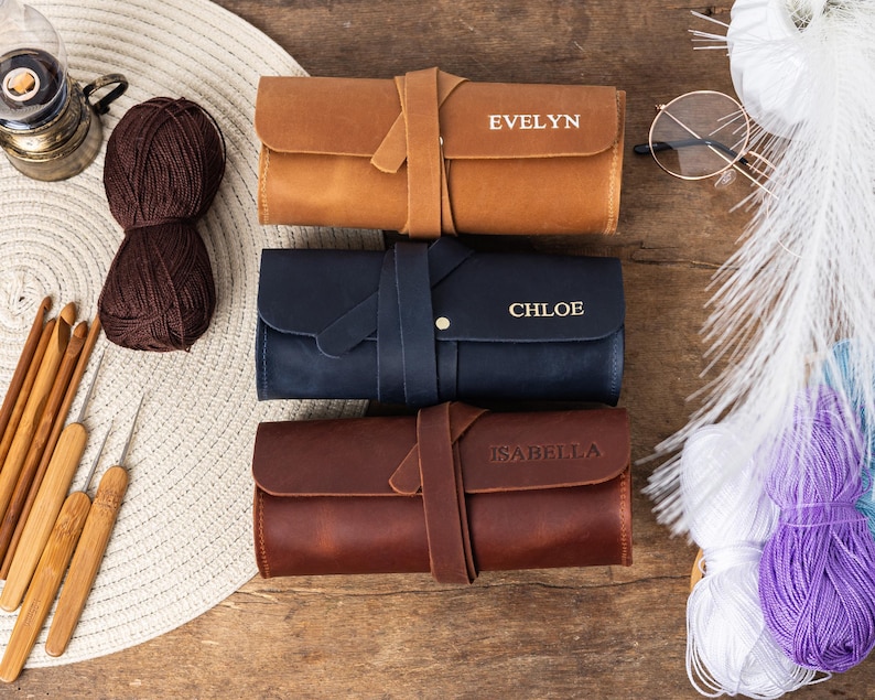 May include: Three brown leather zippered pouches with personalised names: Evelyn, Chloe, and Isabella. The pouches are on a wooden surface with yarn and other craft supplies.